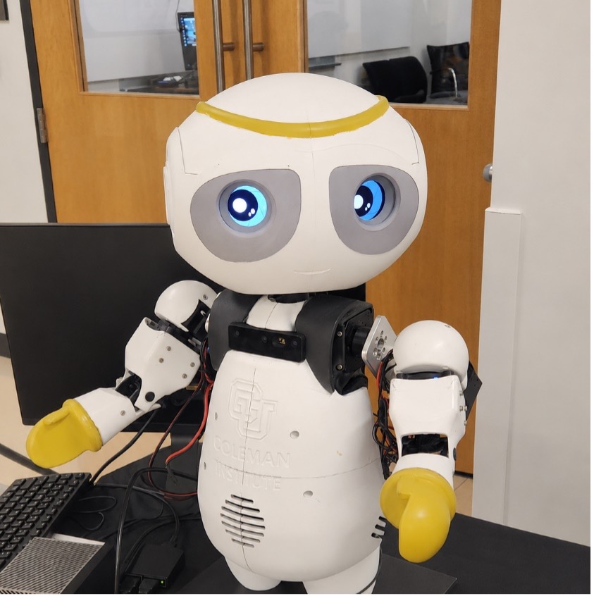 A small friendly robot sits on a desk. C U Coleman Institute Logo on it's chest.