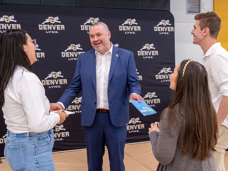 Chancellor Ken Christensen in a blue suit speaks with a small group of students in front of a CU Denver Lynx-branded backdrop.
