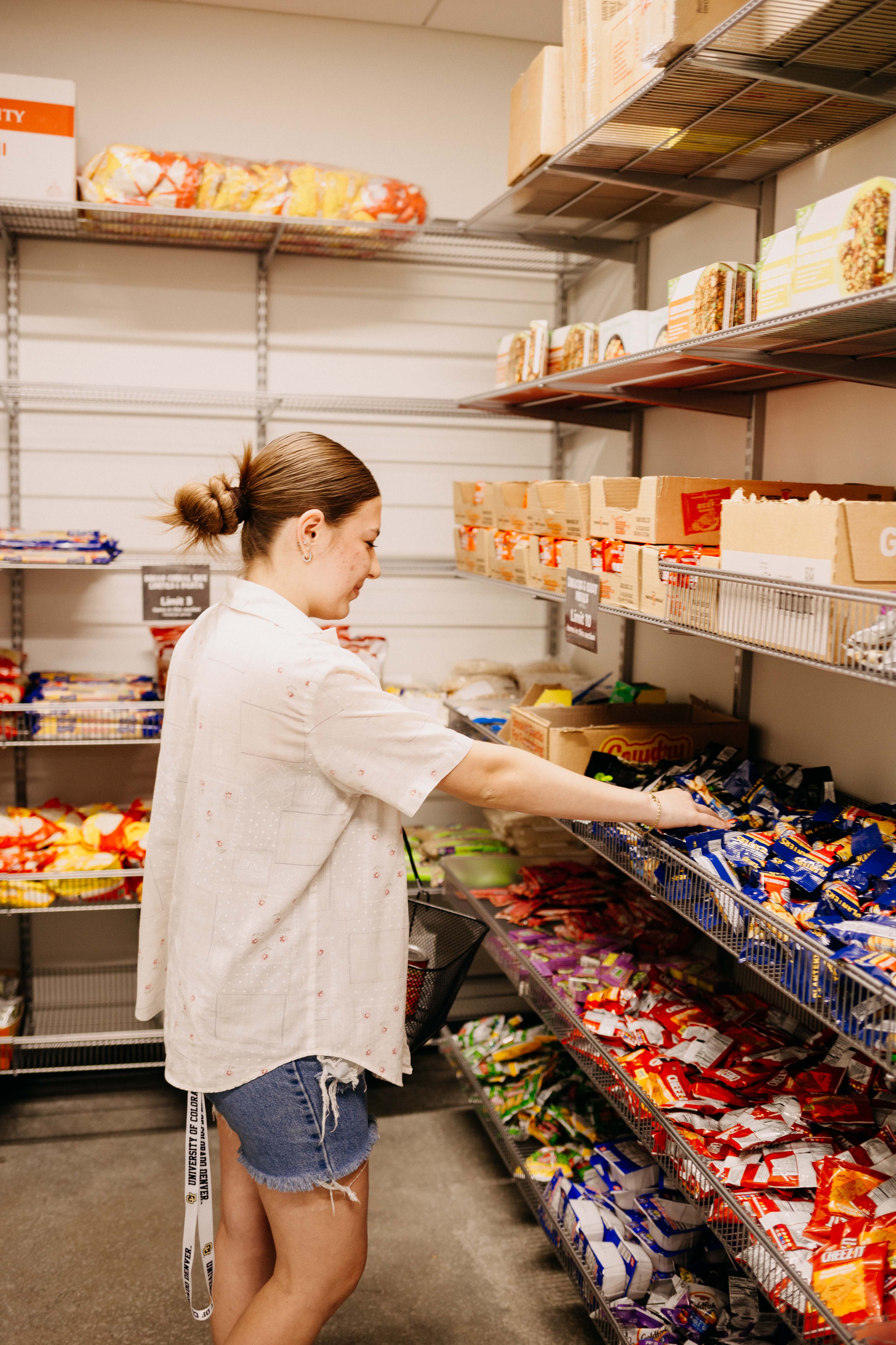 A young woman reaching for a packaged snack on a metal shelf heavily stocked with various food items inside a food pantry.